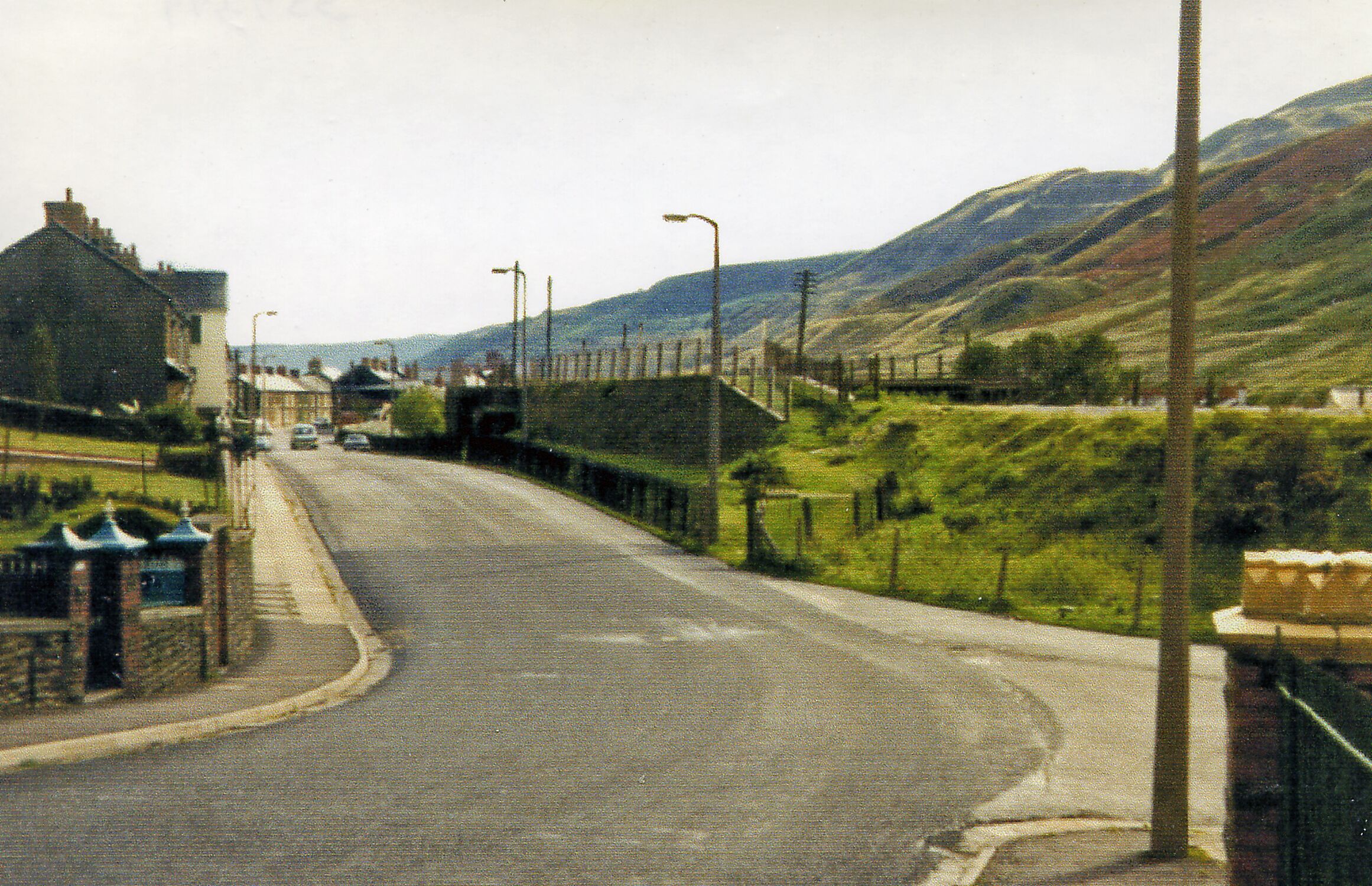 A road in a hilly area with houses on one side and a fence on the other.