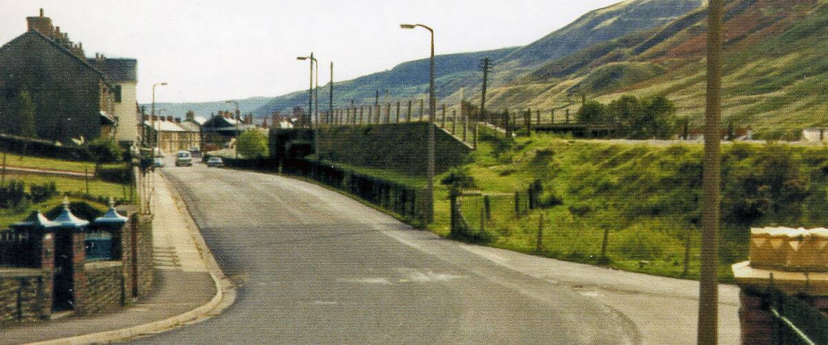 A road in a hilly area with houses on one side and a fence on the other.