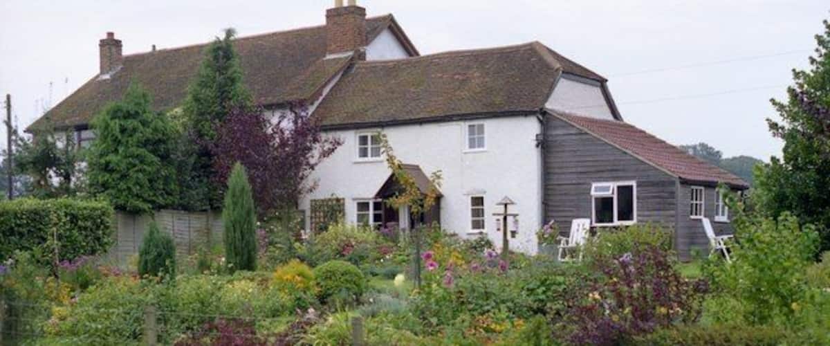 A white house with a grey extension, surrounded by lush gardens and a fence.