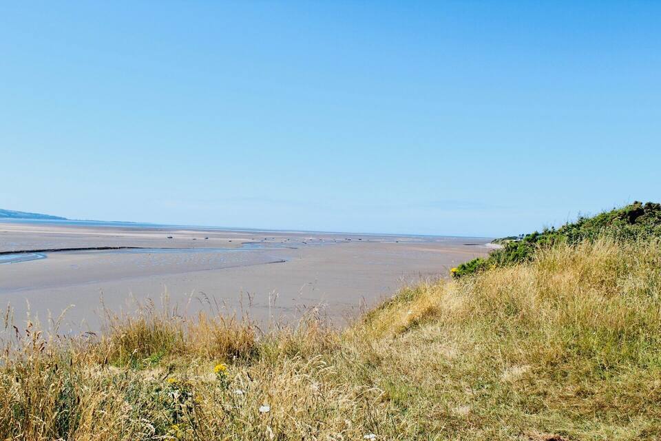 A coastal landscape with a wide sandy beach, grassy dunes, and a clear blue sky.
