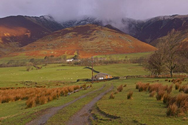 Paisagem rural com uma casa pequena, campos verdes e montanhas ao fundo.