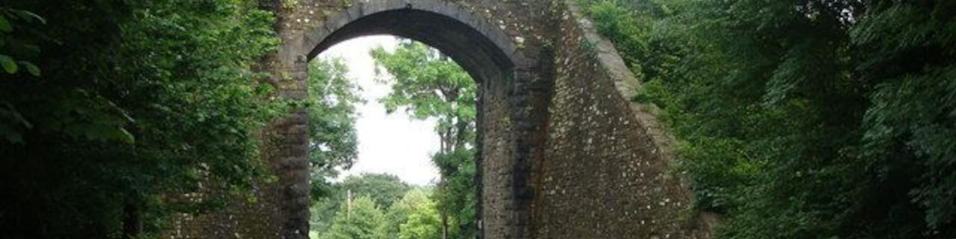 Uma estrada estreita serpenteando sob uma antiga ponte de pedra, em meio a vegetação exuberante.