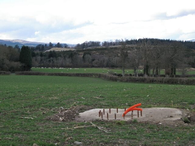 Work in progress by Greycoat Lane Looks like a water point for livestock. Beyond the sheep is a high mound of old clay waste from the Stover Quarry, clothed with gorse and grass. A pile of fresh, light-coloured clay waste appears beyond it, with woodlands and exotic conifers around Stover School showing beyond that.