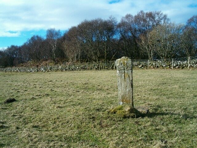 A stone cross in a grassy field with trees and a stone wall in the background.