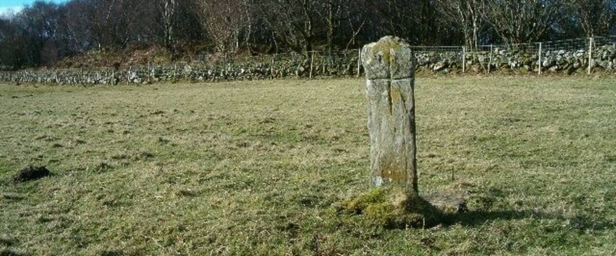 A stone cross in a grassy field with trees and a stone wall in the background.
