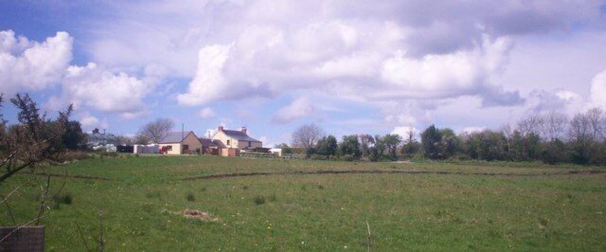 A rural landscape with a green field, a few scattered trees, and a house in the distance under a partly cloudy sky.