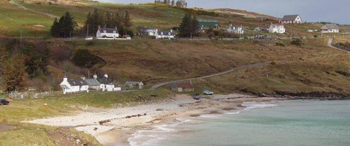 A coastal landscape with a sandy beach, rocky shoreline, grassy hills, and white houses in the background.
