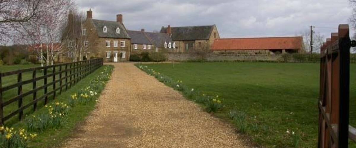 A gravel path leading to a stone building with a red roof, surrounded by fields and a wooden fence.