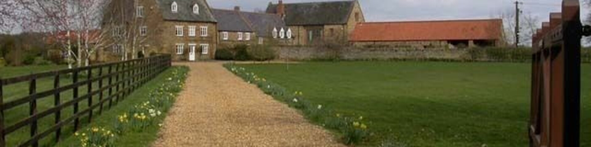 A gravel path leading to a stone building with a red roof, surrounded by fields and a wooden fence.