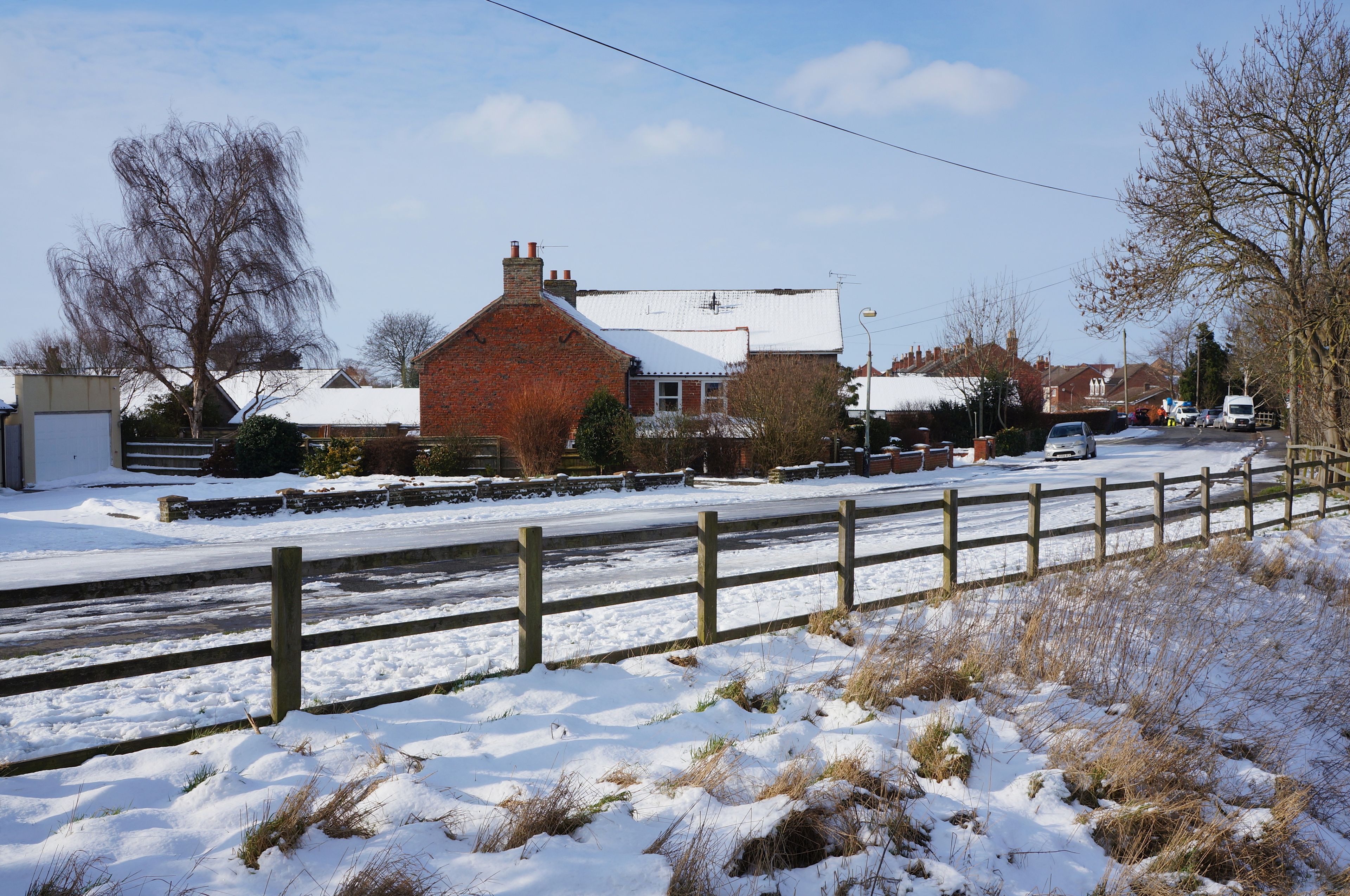 A rural street with snow-covered ground, a brick house, and a wooden fence.