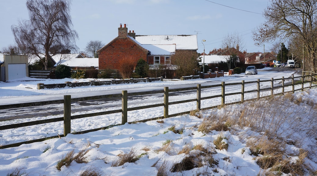 A rural street with snow-covered ground, a brick house, and a wooden fence.