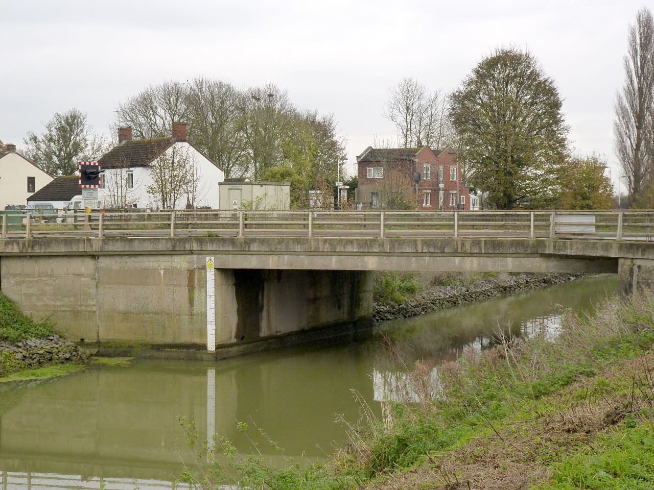 A concrete bridge over a calm river with houses and trees in the background.