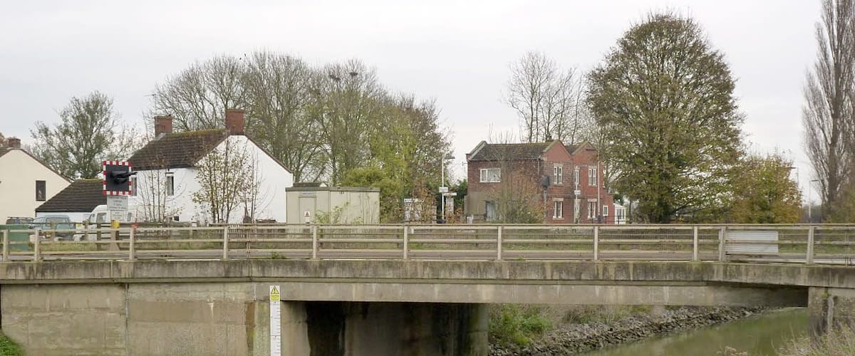 A concrete bridge over a calm river with houses and trees in the background.