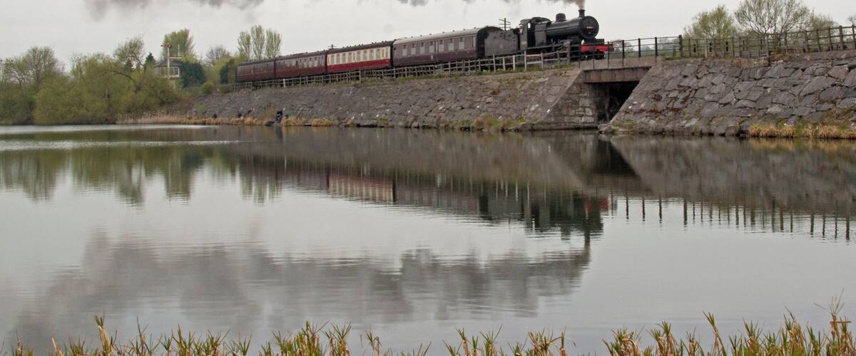 A steam locomotive crossing a bridge over a calm river, with trees and a small building in the background.