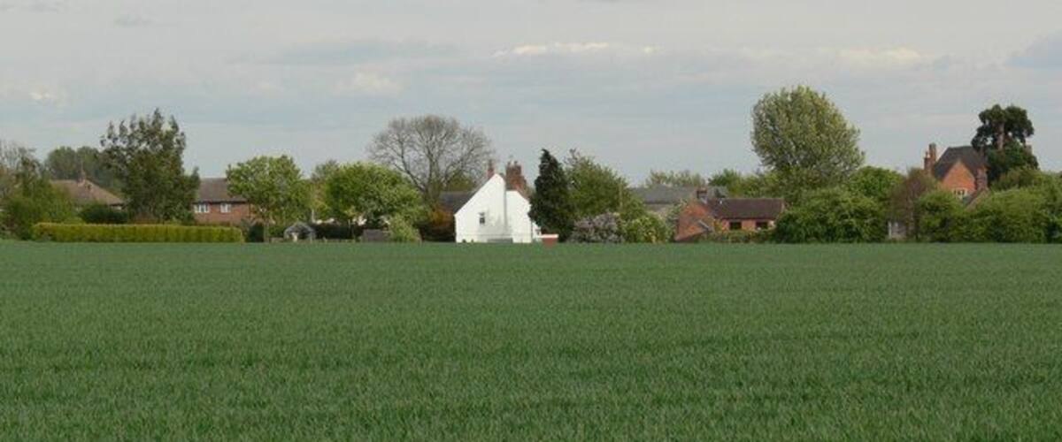 A green field with a white building in the distance, surrounded by houses and trees.