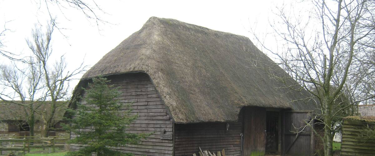 A thatched-roof barn with a dirt path leading to it, surrounded by trees and a grassy field.