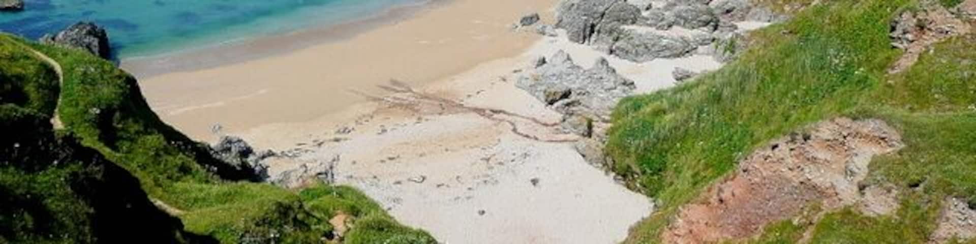 A coastal landscape with a sandy beach, rocky outcrops, and green hills.