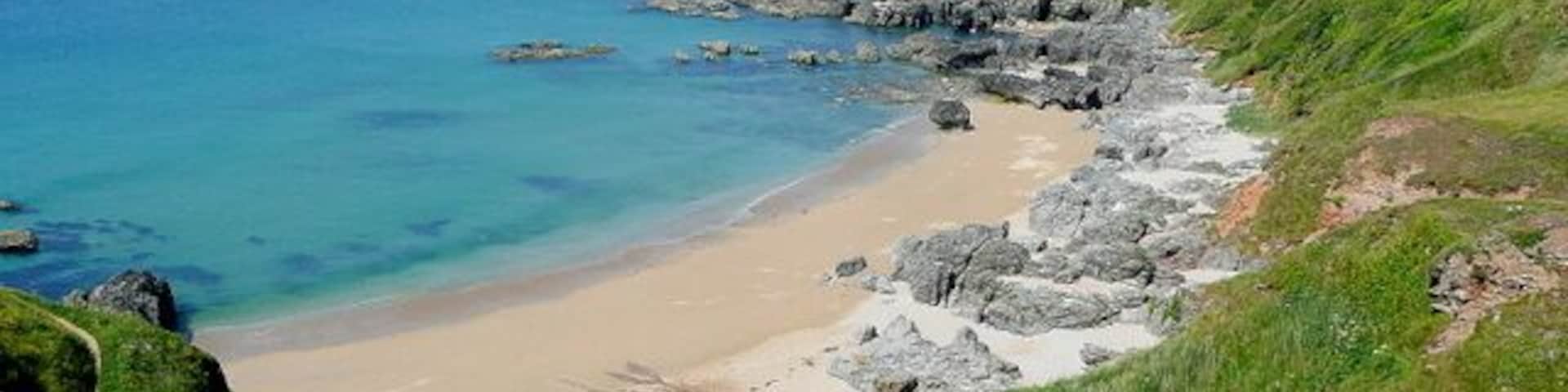 Un paisaje costero con una playa arenosa, afloramientos rocosos y colinas verdes.