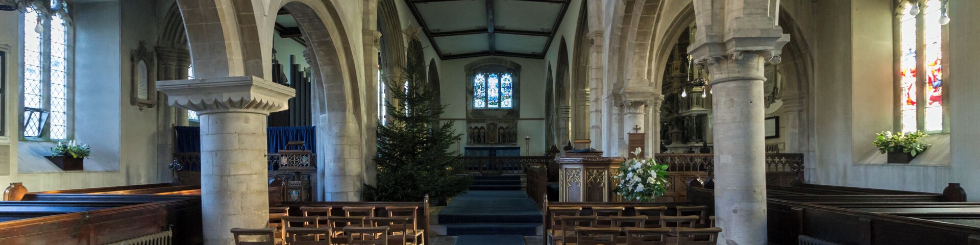 The north arcade is said to date from around 1066 thus it would seem that the church was originally a small Norman church of which only the Nave and Tower remain. The walls and windows of the North and south aisles of the nave date from the Restoration in 1846. Sometime in the 14th century the chancel arch was built, but the chancel it led to has gone. The arch leading to the tower was built about the same time and probably it was then that the nave walls were heightened and a clerestory provided. In 1846 the roof of the chancel was lowered and a low lath and plaster arch constructed under the chancel arch that now exists. This was removed in 1935 and the old arch – sadly hacked about – was revealed. It was impossible to raise the chancel roof, so the space in the apex of the arch was filled by a painting: “Our Lord in Glory”, by Miss Jessie Bayes. In the 15th century, all that now exists beyond the chancel arch and beyond the eastern arches of the nave aisles – was entirely rebuilt. The Turnor chapel is in the north aisle and was built in 1448 by Henry Rochford (The last of that name to live here. He probably built the south aisle rather later). There is a large black and white monument to Sir Edmund Turnor (1619-1707) the founder of the present family of Turnors of Stoke Rochford. Other Turnor memorials are also here. On the floor in the chapel are two medieval monuments. One is the recumbent figure of a knight in armour, and his lady, with a curious covering over them. Probably this monument was in the North stoke church, and after that church was abandoned and ceased to exist, was, after many years exposure in the open, brought here in the 19th century and placed in the middle of the marble floor. No one knows whom the figures represent, though 100 years ago it was thought to be a Neville monument and that name was cut on the surrounding stone work. Bishop Hine believes the arms on the shield to be Cantelupe. The other monument in the North Wall; it is a tomb in a canopied recess, with angels bearing shields, all however, blank today. Nothing is known of this monument. In the south aisle there is the Cholmeley chapel. The most striking object is the large monument erected in 1641 to the memory of Sir Henry Cholmeley (d 1632) the first of the family which still lies at Easton Hall in the parish. The family memorials extend down the south wall including a war memorial to all the Cholmeleys who died in the Great War. Beneath the carpet in the chancel are the Rochford brasses. The Rochfords held Stoke for most of the 15th century. One brass is Henry Rochford, the last of the name, in full armour, who died in 1470. He was the builder of the chapels. The other are his wife with her later husband, Oliver St John. An inscription records that Oliver St John’s mother married eventually the Duke of Somerset, and (through her famous daughter Lady Margaret Beaufort) became grandmother of King Henry VII. The reredos dated 1911 was designed by Mrs G F Watts. The figures represent (from left) St Hugh of Lincoln, St Gilbert of Sempringham, and the Blessed Virgin Mary. Those on the right are St John, Bishop King of Lincoln and St Christopher. The font is probably 1846 but the cover designed and given by Mr C H Turnor was placed here in 1937. It follows in feeling the Jacobean work of the 17th century. The stone work of the tower has deeply recessed windows with a Norman string course beneath the belfry windows. The balusters in these windows, one with a criss-cross early date – possible Saxon – and Transitional Norman. The battlemented top and the west window were doubtless added in the 15th century. (From guide book.)