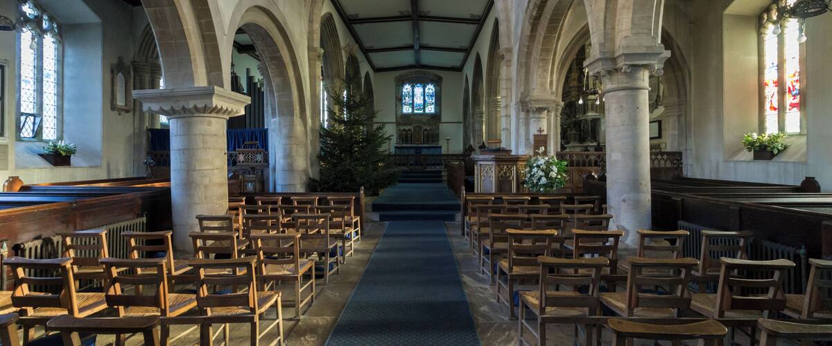 A church interior with wooden pews, a central aisle, stained glass windows, and a vaulted ceiling.