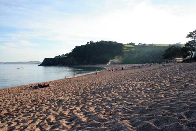 Evening on Blackpool Sands. Looking westwards across the bay with the dead calm sea reflecting the early evening sun. The main road to Kingsbridge follows the coast here and can just be made out through the plume of smoke. The land on the horizon is at the other end of Start Bay with Start point just out of shot on the left, see 574819.