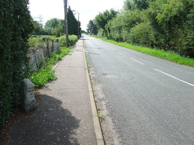 A rural road with a sidewalk, greenery, and a stone marker.