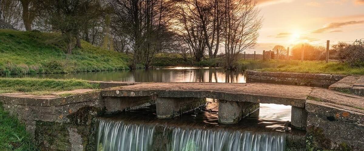 A waterfall over a stone bridge, surrounded by trees and a river.