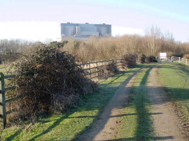 A rural dirt road with a fence on one side and a building in the background.