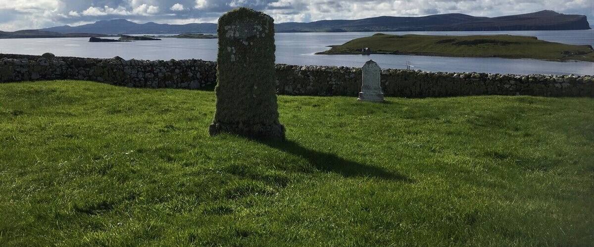 A grassy field with a stone monument and a smaller headstone, overlooking a body of water and distant hills.