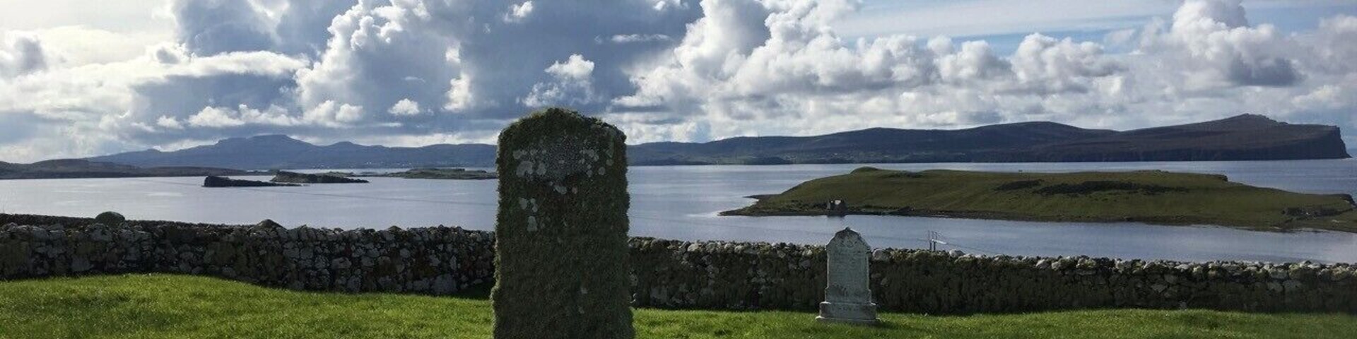 A grassy field with a stone monument and a smaller headstone, overlooking a body of water and distant hills.