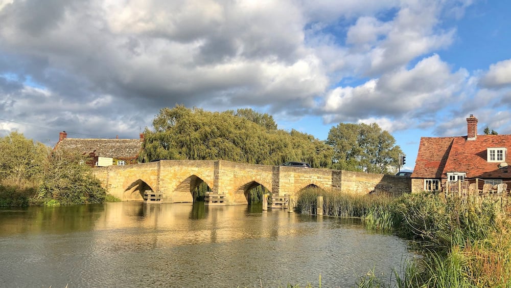 A stone bridge over a calm river, surrounded by lush greenery and a partly cloudy sky.