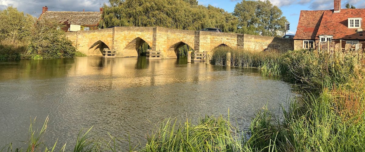 A stone bridge over a calm river, surrounded by lush greenery and a partly cloudy sky.