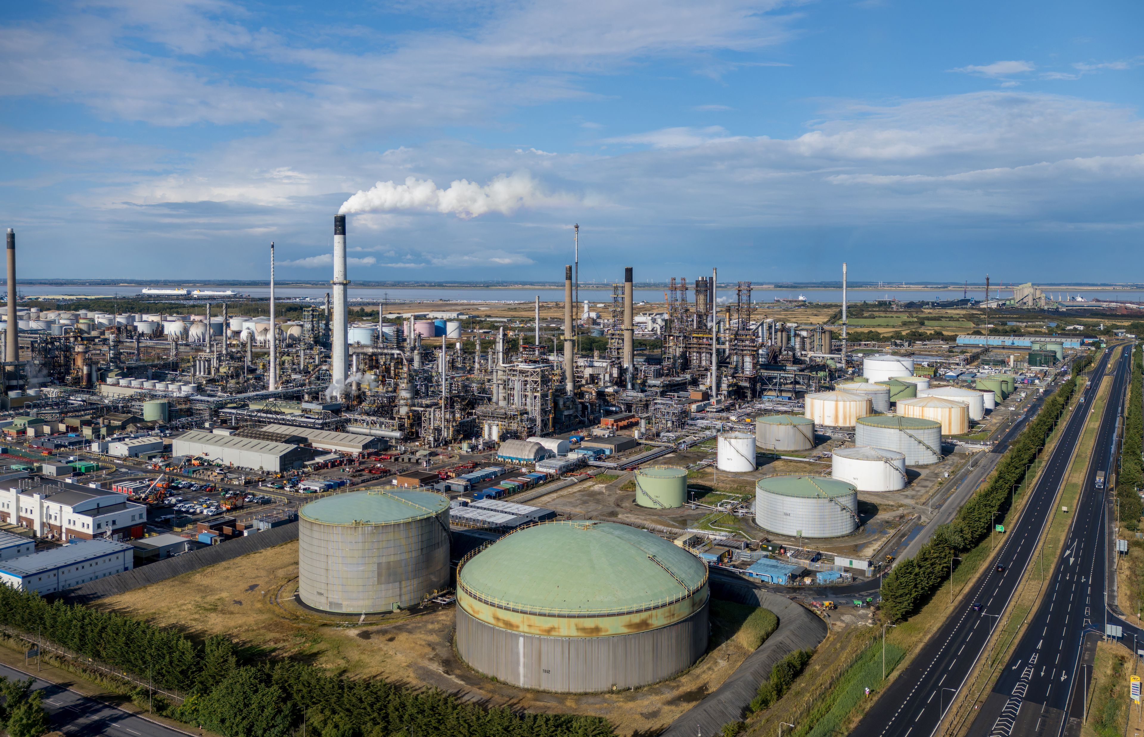 An industrial complex with multiple storage tanks, tall chimneys, and a highway running alongside.