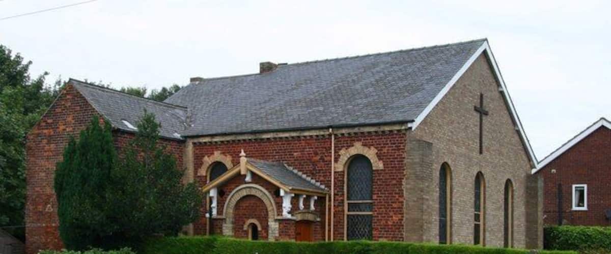A brick church with a cross on the front, a bell tower, and a small shed in the foreground.