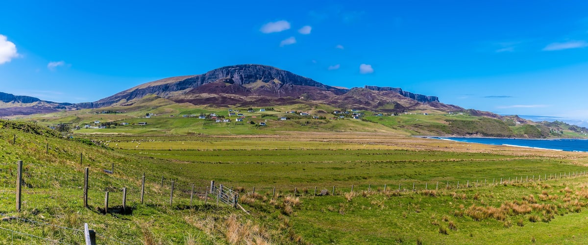 A coastal landscape with a prominent mountain, green fields, and a small village.