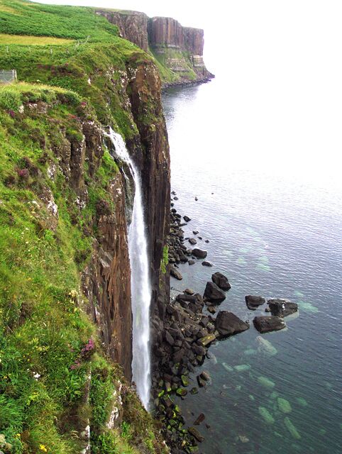 A coastal cliff with a waterfall, rocky shoreline, and green vegetation.
