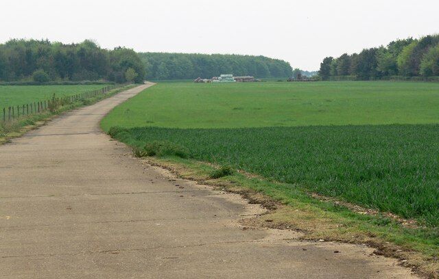 A rural road curving through green fields with a fence on one side and a building in the distance.