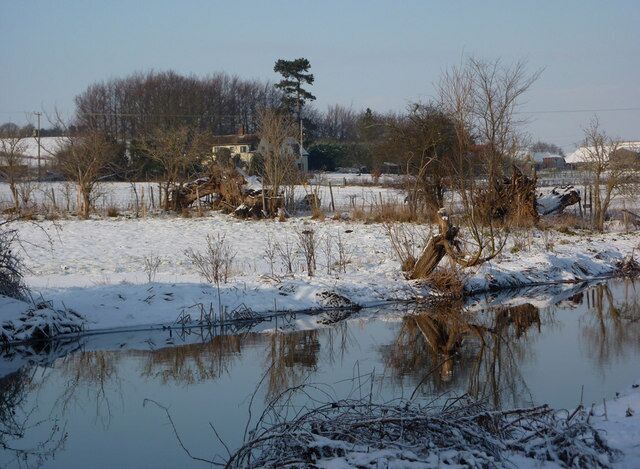 A snow-covered landscape with a frozen pond, bare trees, and a distant building.