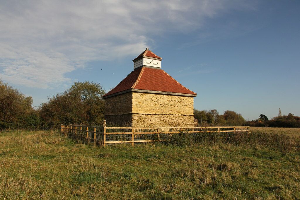 A stone structure with a red roof and a wooden fence in a grassy field.