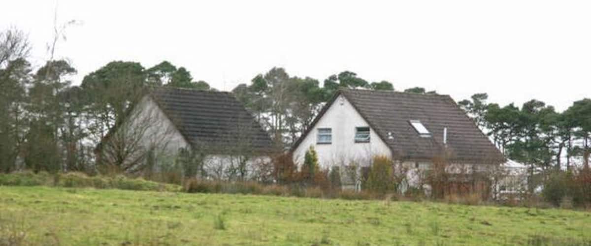 Two white houses with grey roofs in a grassy field, surrounded by trees.