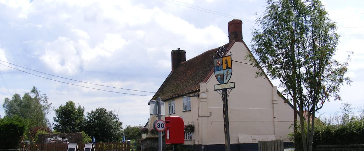 A rural road with a speed limit sign, a pub named 'The Swan' with a traditional white facade, and a signpost with a coat of arms.