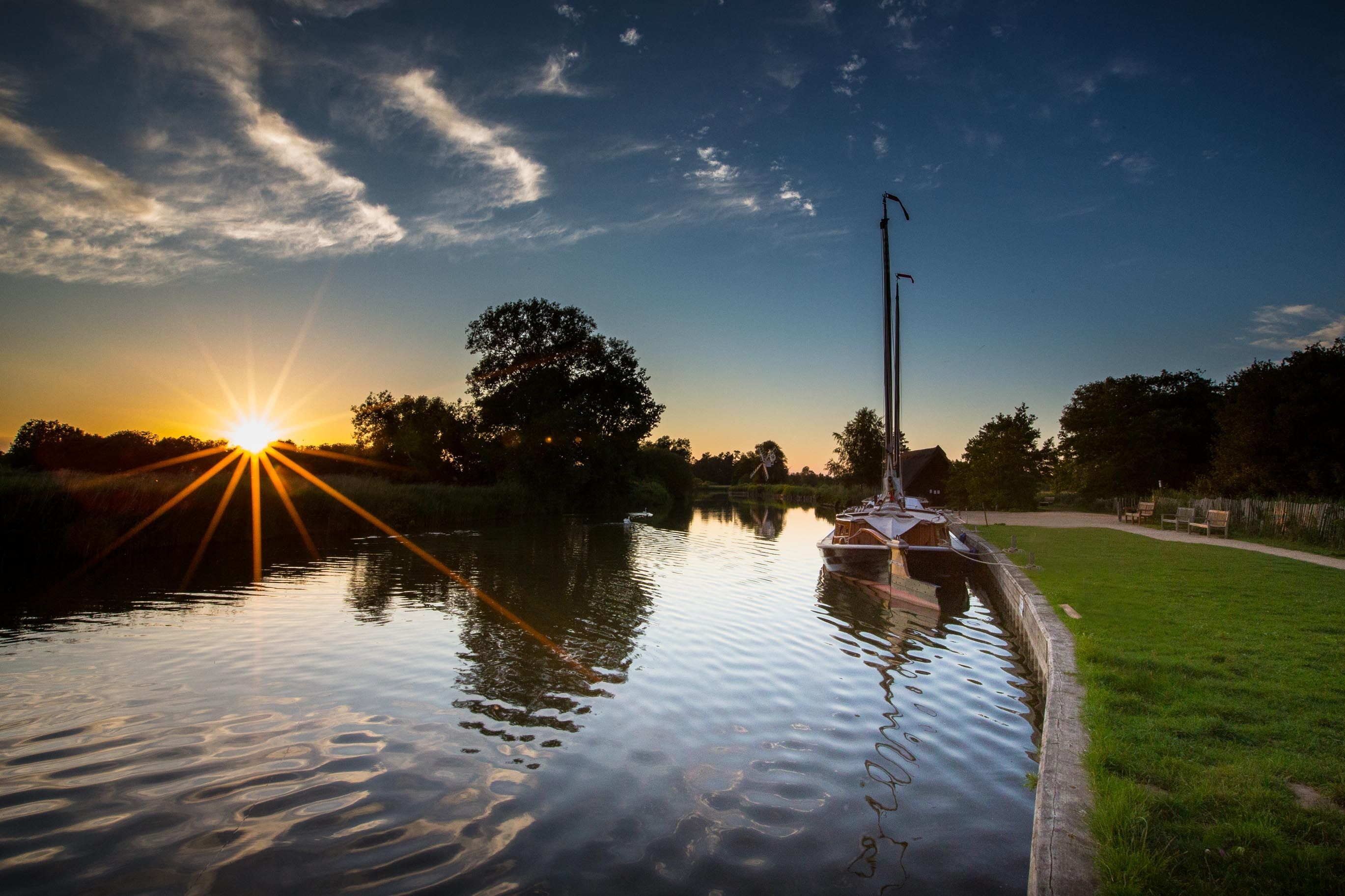 A calm river at sunset with a boat docked on the right, surrounded by trees and a grassy bank.