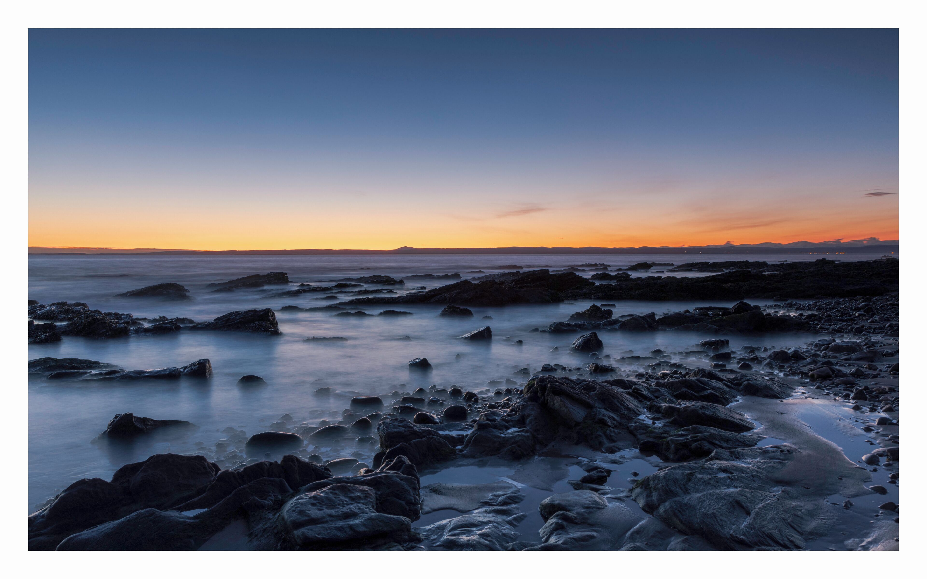A rocky shoreline at sunset with a calm sea and a gradient sky.