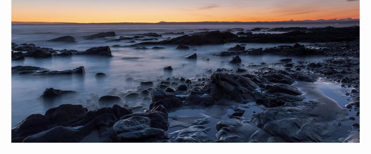 A rocky shoreline at sunset with a calm sea and a gradient sky.