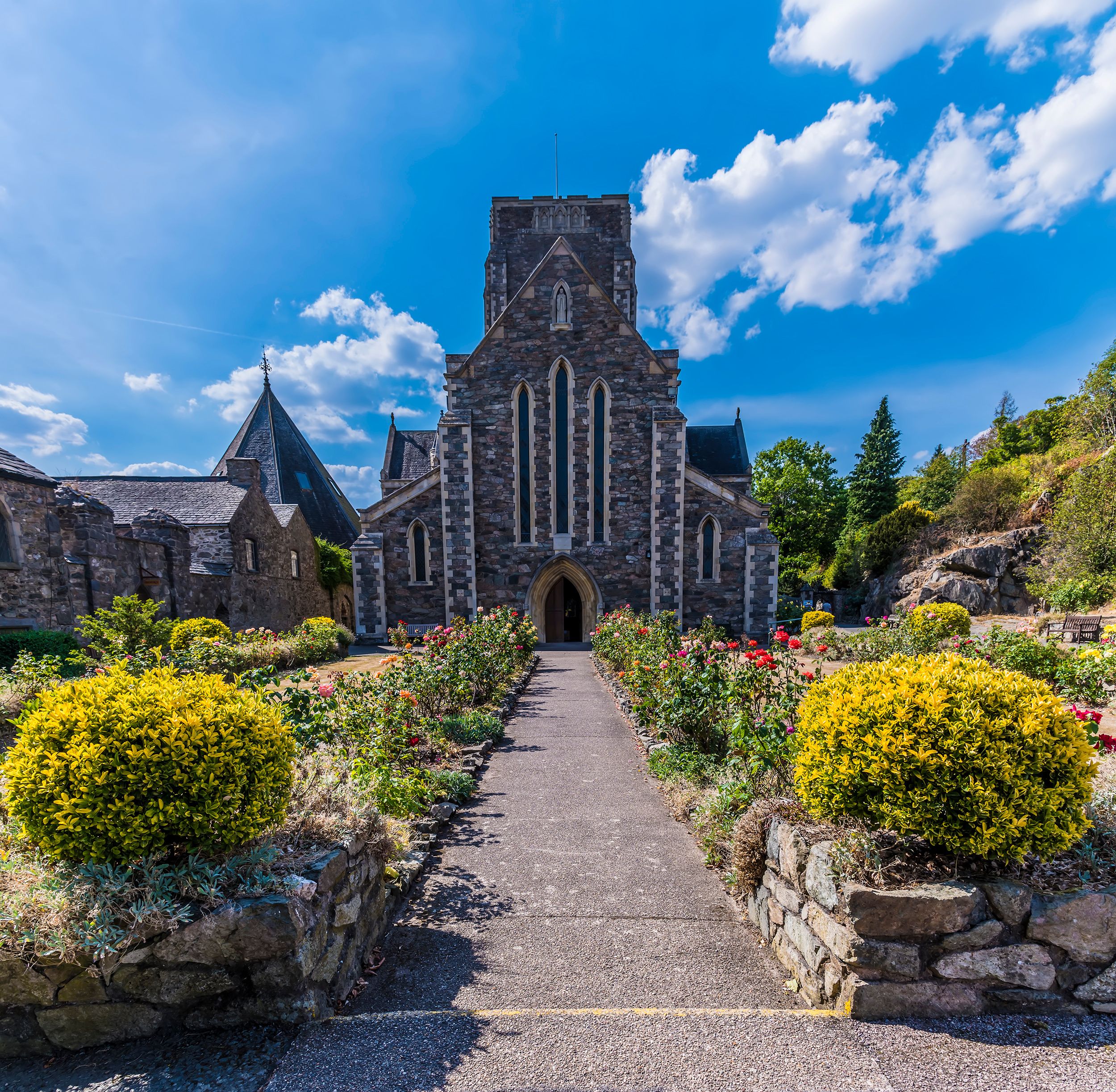 A historic stone church with a pointed steeple, surrounded by well-maintained flower beds and a clear blue sky.