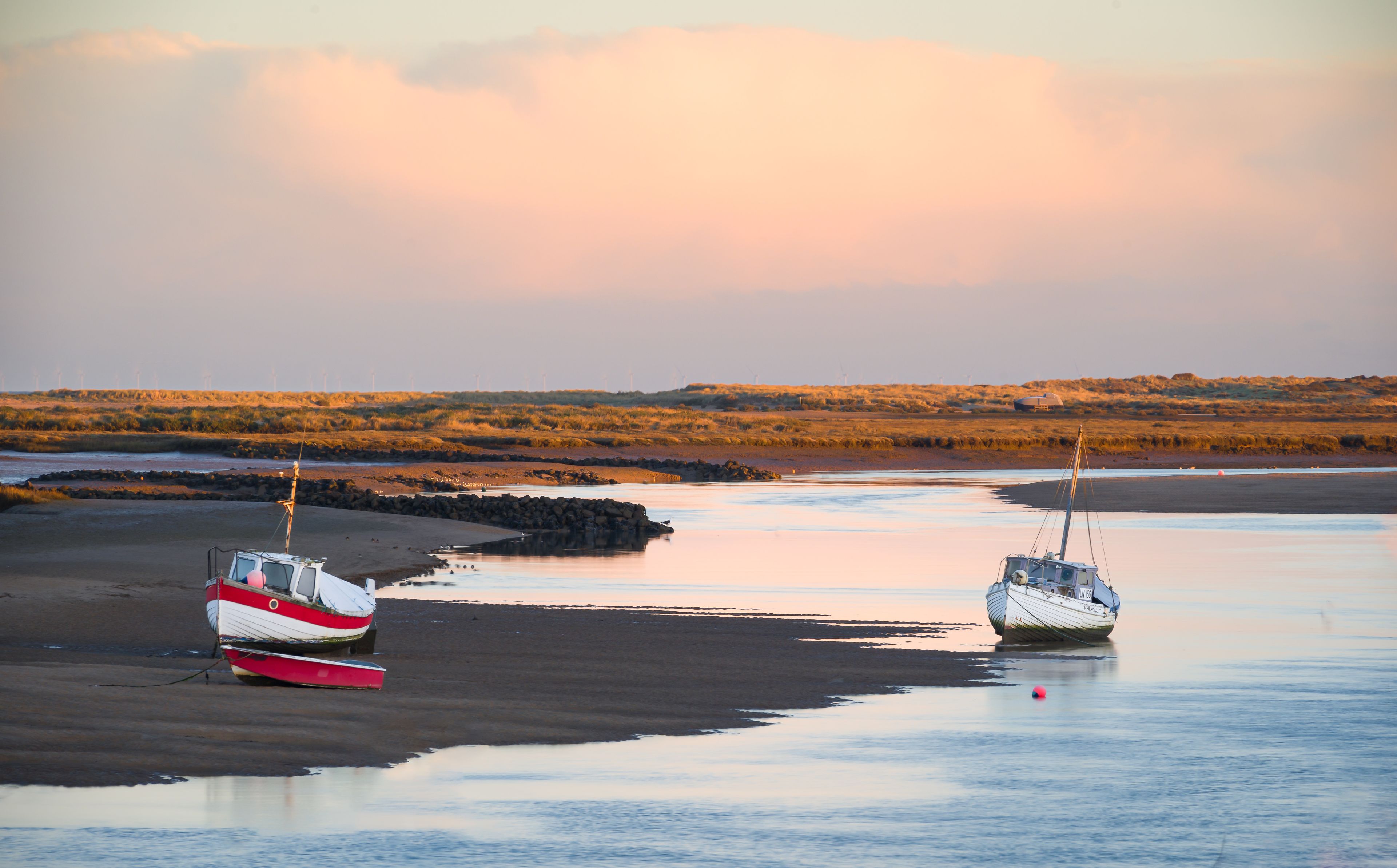 Dois barcos em uma margem de rio calma, com paisagem gramada e turbinas eólicas ao longe.