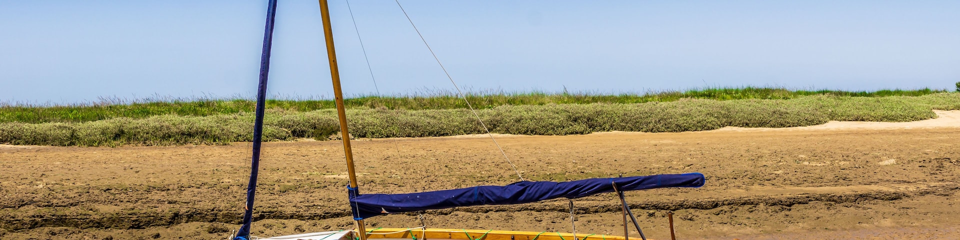 Um pequeno veleiro chamado 'Zither' com toldo azul, ancorado perto de uma praia arenosa com grama ao fundo.