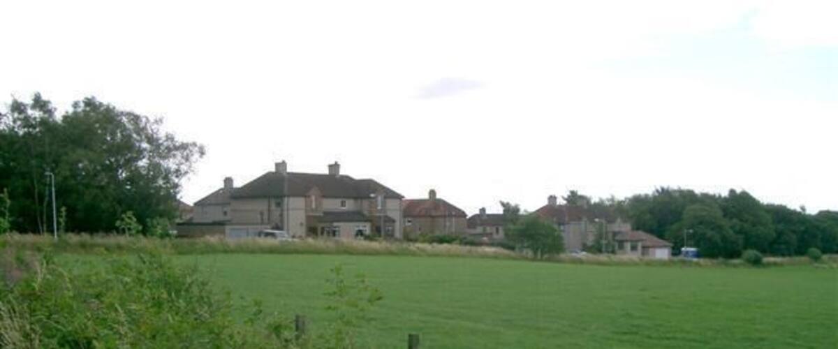 A large green field with a fence in the foreground and a row of houses in the background.