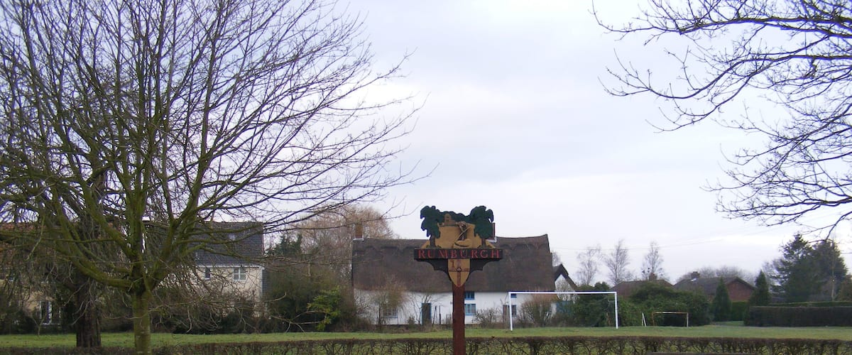 A signpost with 'Kimbell' written on it, a bench, a hedge, and a house in the background.