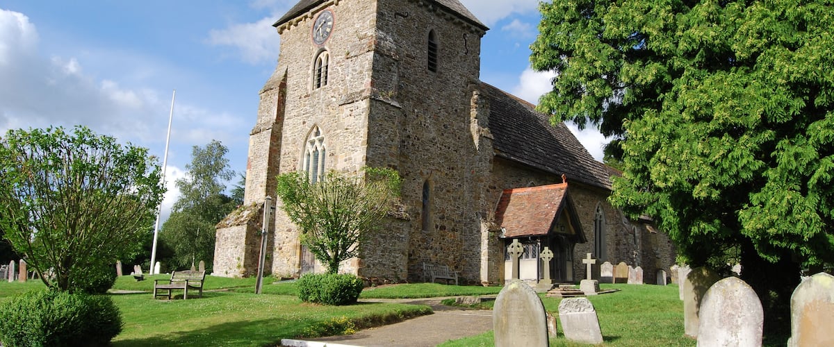 A historic stone church with a clock tower, surrounded by a cemetery with headstones and a well-maintained lawn.